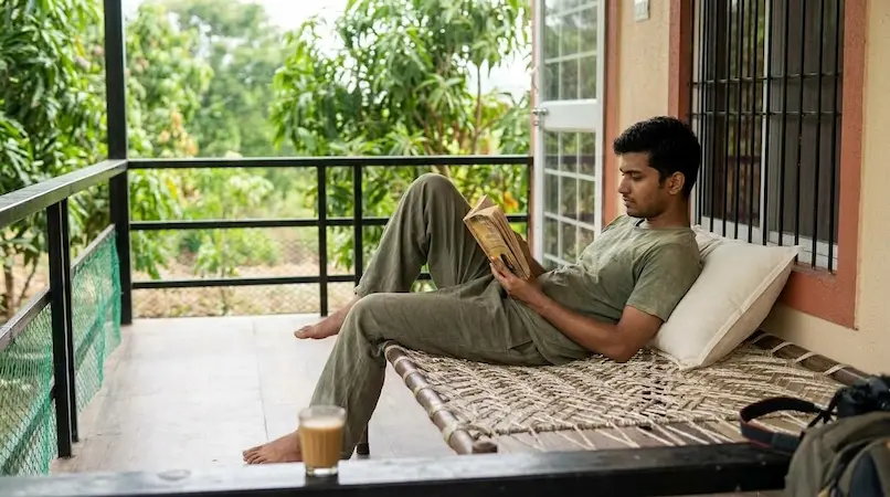 Guest relaxing on wooden charpai cot reading a book with cutting chai on private porch gallery at Bimal Farm